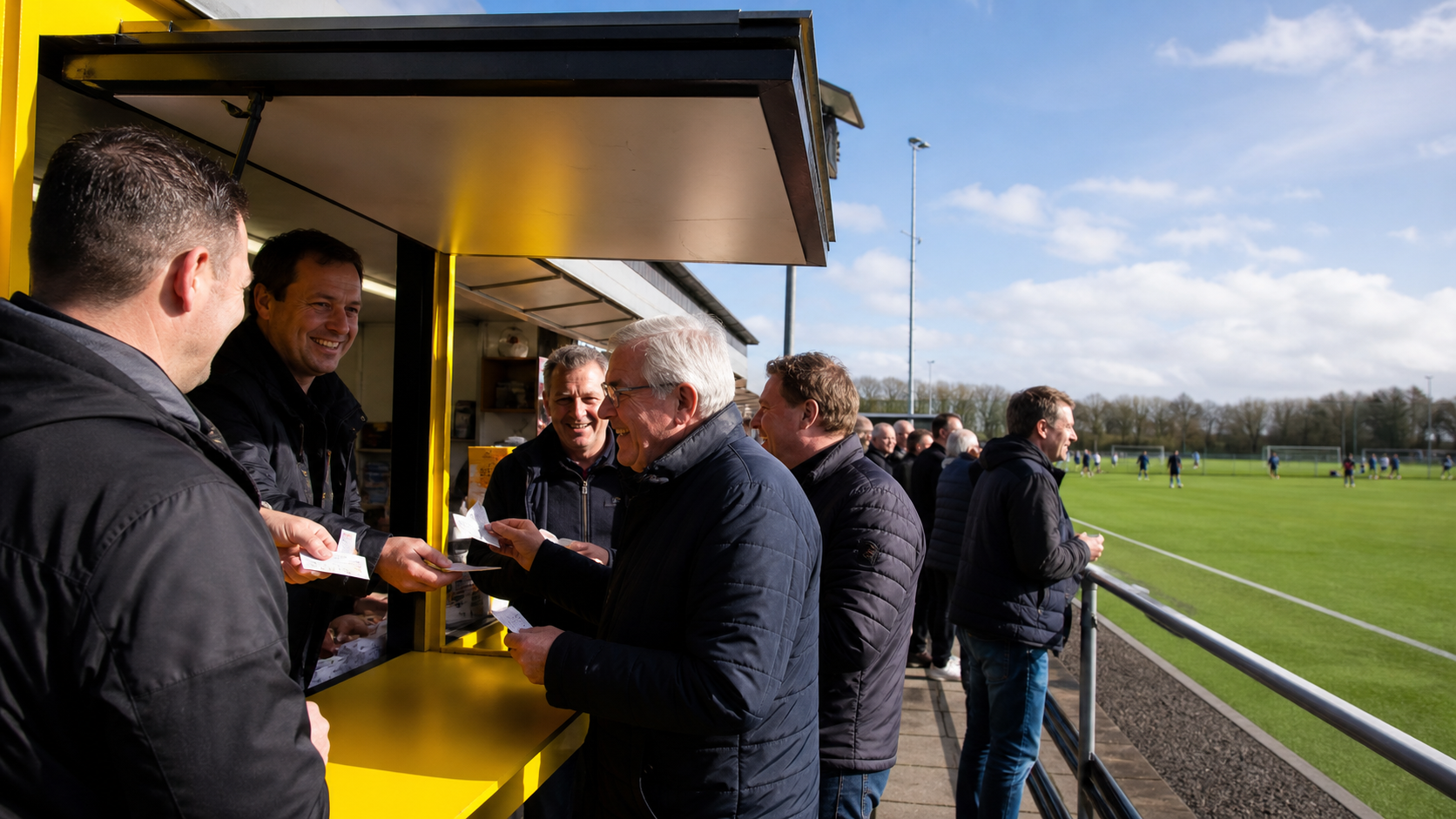 Supporters kopen loten bij een geel loket naast het voetbalveld